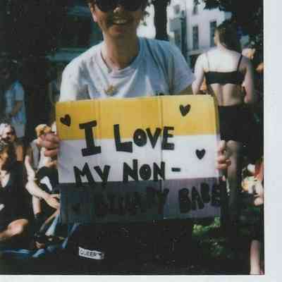A person wearing sunglasses holds a sign reading, "I love my non-binary babe".