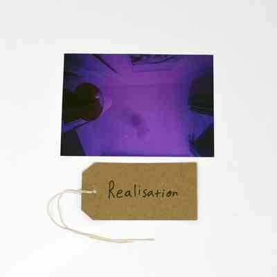 A purple photo of a ceiling with a brown handwritten luggage tag inscribed with "Realisation"