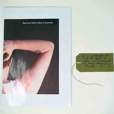 Booklet entitled "Teach me what it takes to be proud." The cover image is a close up of someone raising their right arm, taken from behind. Also a brown, handwritten luggage tag.