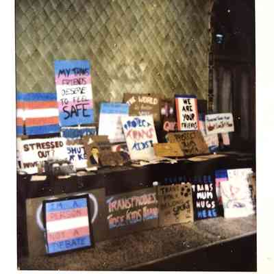 The photograph shows a collection of signs advocating for trans rights, displayed on a surface.