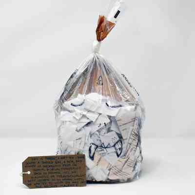 A clear plastic bread bag filled with ripped up paper documents and knotted. A cartoon face of a monster has been drawn on the bag. With a brown, handwritten luggage tag.