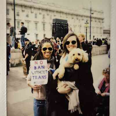 Two smiling individuals hold a small dog and a sign reading "No Ban Without Trans" during a public protest.
