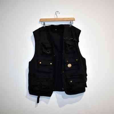 A black gilet with several pockets on a wooden clip hanger. It has a yellow badge (with a cat in the trans colours on it) pinned to the left breast pocket and is held together near the left shoulder with safety pins