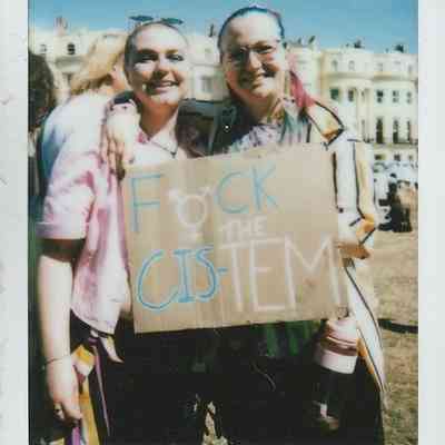 Two people at a protest are holding a cardboard sign which says "Fuck the Cis-Tem".