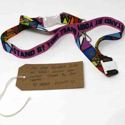 A colorful lanyard with “STAND BY YOUR TRANS” printed on it, with a brown luggage tag.