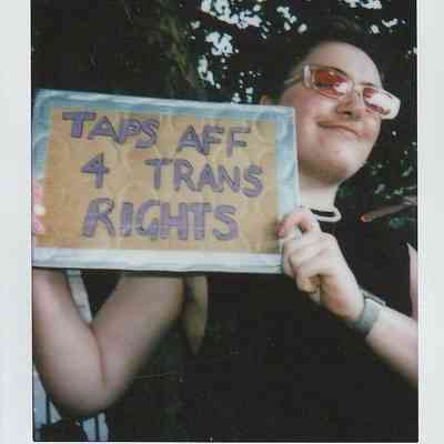 A person is holding a sign that reads "Taps Aff 4 Trans Rights" while smiling confidently