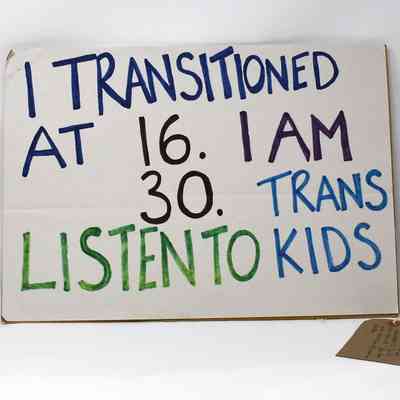 A white, cardboard placard with the text "I TRANSITIONED AT 16. I AM 30. LISTEN TO TRANS KIDS" in blue, black and green. With a brown, handwritten luggage tag.