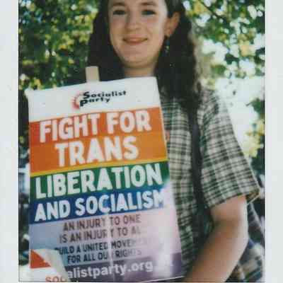 A person holds a colorful sign advocating for trans liberation and socialism at an outdoor event beneath trees.