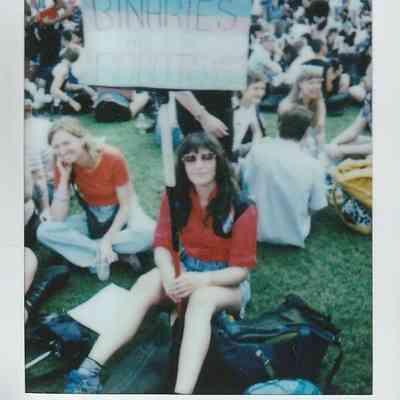 A group of people sits on grass at a protest holding a sign that reads "Binaries Are For Robots".