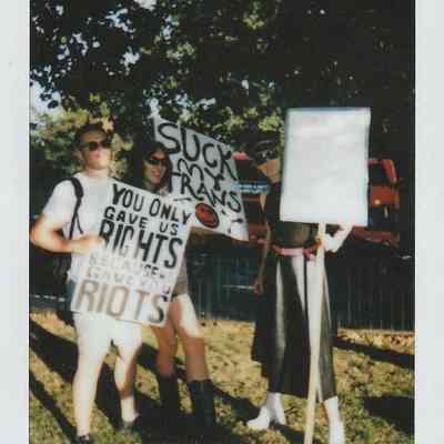 Three people stand holding protest signs in a sunlit park setting, holding placards.