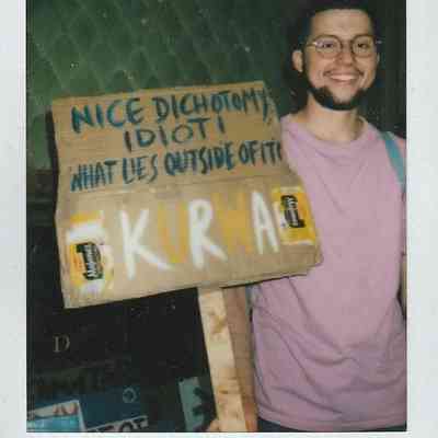 A smiling person holds a cardboard sign with various messages, standing against a textured background in TP Brighton.