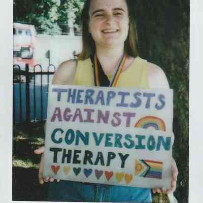 A person smiles while holding a sign stating "Therapists Against Conversion Therapy" with colorful decorations.