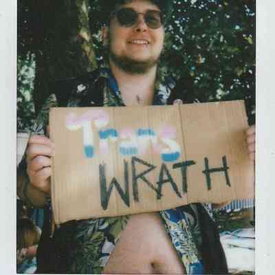A person smiles while holding a sign saying "Trans WRATH" in a sunny outdoor setting.