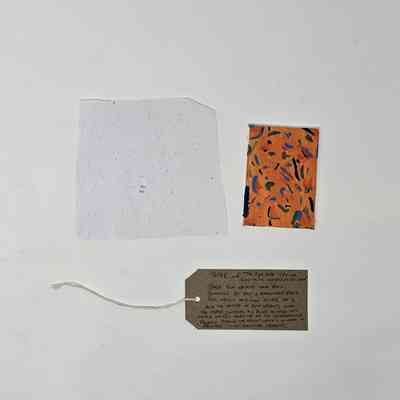 A white handmade piece of paper, a small red painting and a brown tag with writing on it lay on a white surface.
