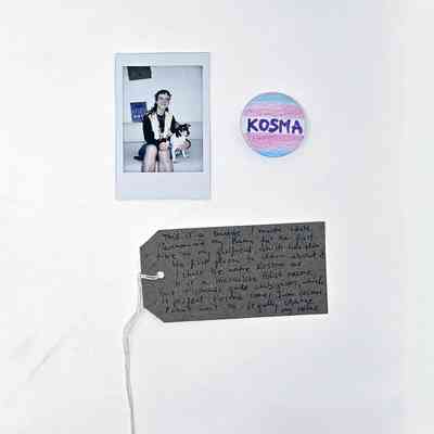 A small badge coloured in the trans flag colours and featuring the name "KOSMA". With a Polaroid of the donor and a brown, handwritten luggage tag.