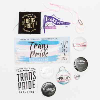 The image features a collection of badges, keychains, and informational badges promoting Trans Pride Brighton, with dates detailed for July 25th, 26th, and 27th. Various designs include phrases and graphics highlighting the theme, such as "Putting the 'T' First," and "Brighton Transformed," signifying support and celebration for the transgender community. The badges display different variations of the Trans Pride logo from multiple years, reflecting community engagement and awareness over time in Brighton and Hove.