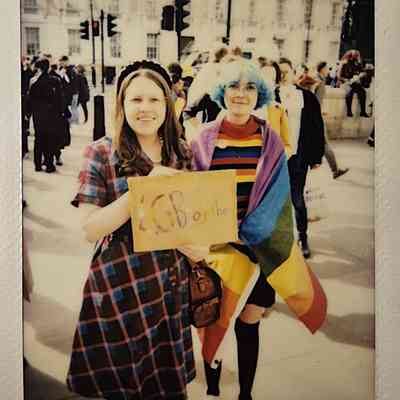Two people stand smiling, one has a rainbow flag wrapped around them and the other is holding a small placard which says 'LGBTOGETHER'.