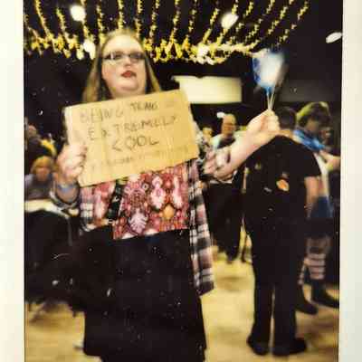 A person holds a sign reading "Being trans is extremely cool" in a lively, festive event space.