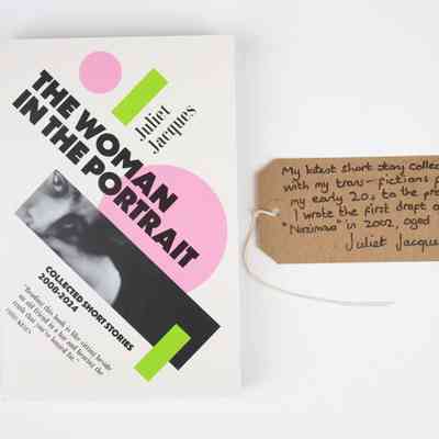 A book titled "The Woman in the Portrait - Collected Short Stories 2008-2024" by Juliet Jacques. The cover is white with green and pink geometric shapes and black and white close up photo of a person's eyes. With a brown, handwritten luggage tag.