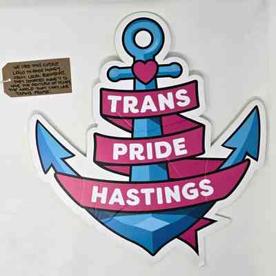 Image of a foam board cut out of the Trans Pride Hastings logo. The logo is a blue anchor with a pink heart a the top and the words "Trans Pride Hastings" in white on a pink ribbon entwined round the anchor. With a brown, handwritten luggage tag.