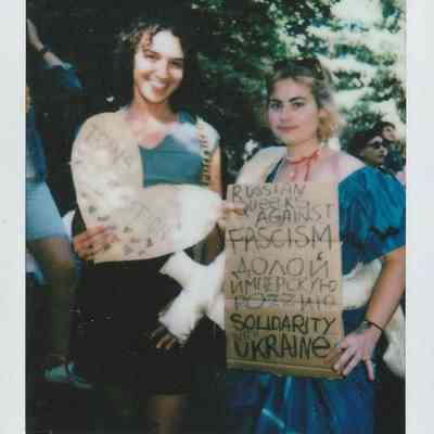 Two people holding signs, one which says "Trans Liberation" and the other "Russian queers against fascism solidarity with Ukraine".