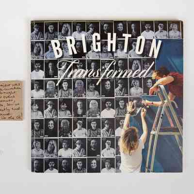 Cover of the paperback book "Brighton Trans*formed." The background is a grid of black and white portraits - to the side there is someone on a ladder putting up one of the photos in the grid, and someone else to the left side of the ladder helping hold the photo up.