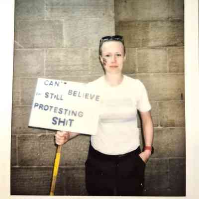 A person stands holding a sign reading, “Can’t believe I’m still protesting this shit,".