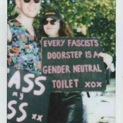 Two people holding a handmade sign with a message stand together outdoors, surrounded by greenery.