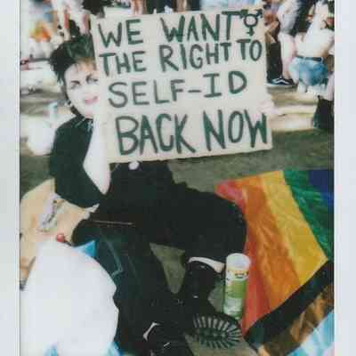 A person sits on the ground holding a sign which says "WE WANT THE RIGHT TO SELF-ID BACK NOW"