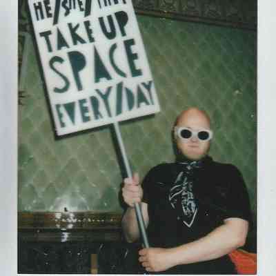 A person holding a sign advocating inclusivity stands confidently in a room, wearing sunglasses and a patterned scarf.