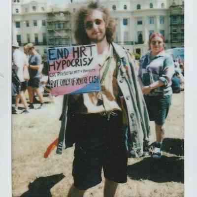 A person stands on grass, holding a sign about "END HRT hypocrisy".