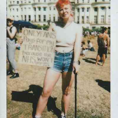 A person stands outside with a cane, holding a sign which says "Only Fans Pays For My Transition Because The NHS Won't".