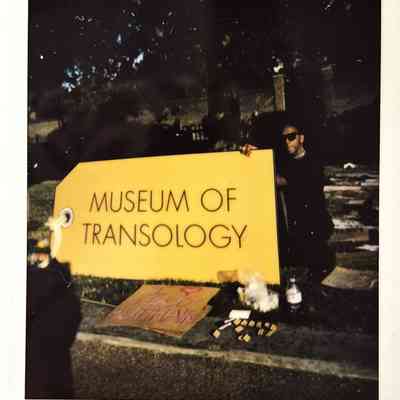 A person poses beside a large, yellow sign labeled "Museum of Transology," with various items nearby.