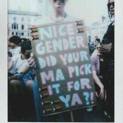 A person smiles, holding a sign at London Trans Pride which says "Nice Gender Did Your Ma Pick It For Ya?!"