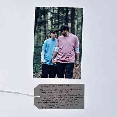 A photo of two people holding hands, and each others gaze, in a wood celebrating their engagement. Both wear baseball caps, one a blue t-shirt, the other a pink t-shirt. With a brown handwritten luggage tag.