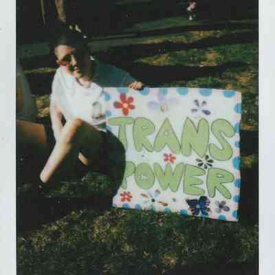 A person is sitting on grass, holding a colorful sign that says “Trans Power” in large letters.