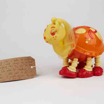 A children's toy which is a ladybug that is yellow and has an orange/red shell. It has hair on it almost like a my little pony. There is also a brown luggage tag  with writing on.