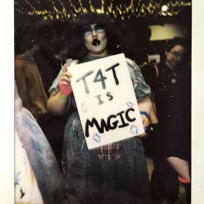 A person holds a sign saying “T4T is magic” with colorful decorations underneath vibrant lights.