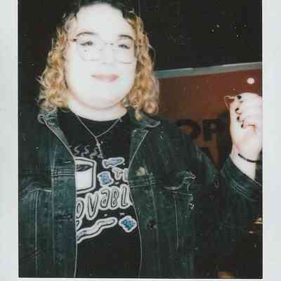 A person with curly hair is smiling, wearing glasses, and holding a necklace card in indoor setting.