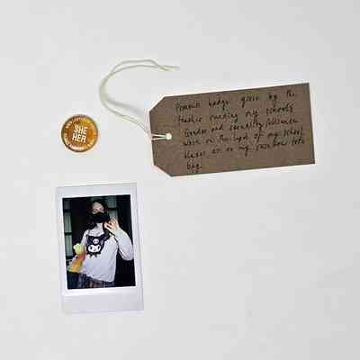 A small, orange, "SHE HER" pronoun pin badge from LGBTYOUTH. With a Polaroid and a brown, handwritten luggage tag.