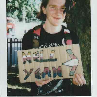 A person smiles while holding a colorful sign saying "HELL YEAH," wearing a floral crown.