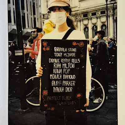A person wearing a mask holds a sign listing names then "PROTECT BLACK TRANS LIVES".