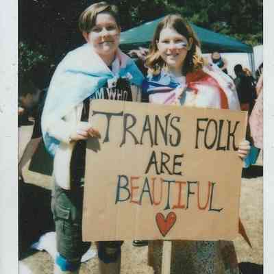 Two people are holding a sign reading "Trans Folk Are Beautiful" with flags draped around them.
