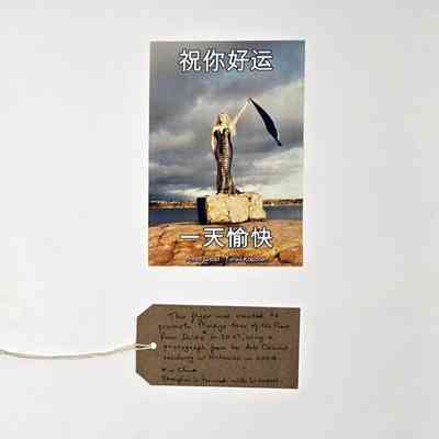 Promotional postcard for Mandy Romero's tour of China. There is Chinese text at the top and bottom and the image is of a person in a dress standing on a rock holding a scarf up in their left hand. With a brown, handwritten luggage tag.