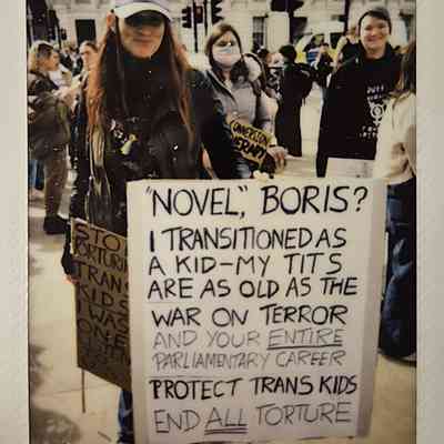 A protester holds a sign which says "NOVEL, BORIS? I TRANSITIONED AS A KID-MY TITS ARE AS OLD AS THE WAR ON TERROR AND YOUR ENTIRE PARLIAMENTARY CAREER PROTECT TRANS KIDS END ALL TORTURE".