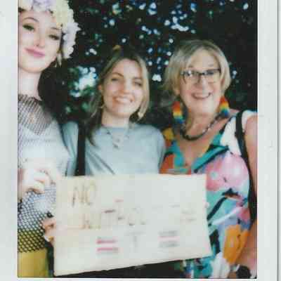 Three smiling individuals pose together, one holding a sign, with trees blurred in the background.