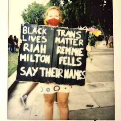 A person wearing a red mask holds a sign advocating Black and Trans Lives Matter.