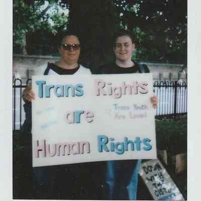 Two individuals hold a sign reading "Trans Rights are Human Rights" during a gathering outdoors.