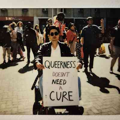 A person holds a sign saying "Queerness doesn't need a cure" whilst they are sat in their wheelchair.