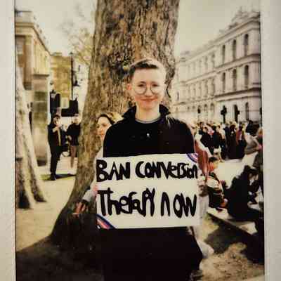 A person holding a sign reading "Ban Conversion Therapy Now" stands near a large tree.